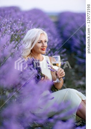 Blonde lavender field holds a glass of white wine in her hands. Happy woman in white dress enjoys lavender field picnic holding a large bouquet of lavender in her hands . Illustrating woman's picnic 129771598