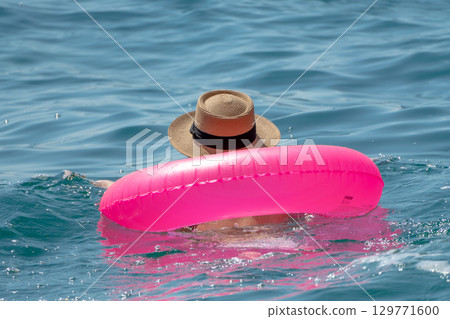 Swimmer, Floating, Ocean: Person relaxing with bright pink inflatable ring and straw hat in calm blue sea on summer day. Swimmer, Floating, Ocean: Person relaxing with bright pink inflatable ring and straw hat in calm blue sea on summer day. 129771600