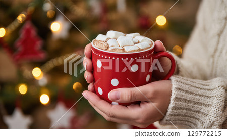 Christmas cocoa with marshmallows in red mug held by woman in cozy sweater near festive decorated tree. Warm holiday vibes. 129772215