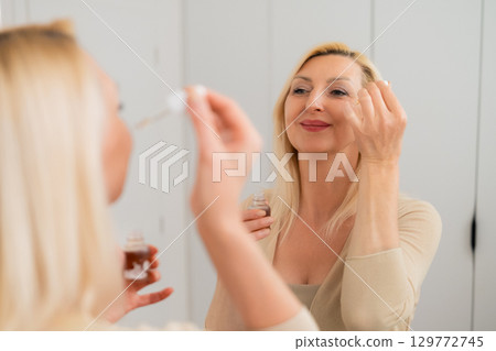 Stylish blonde lady in nude clothing using facial serum to moisturize and refresh skin while standing by mirror indoors Stylish blonde lady in nude clothing using facial serum to moisturize and refresh skin while standing by mirror indoors 129772745