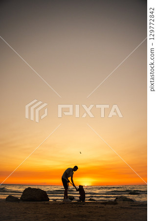 Man playing with his German Boxer dog on the beach at sunset, holding the dog's paw while standing by the ocean waves, silhouette against the colorful sky. 129772842