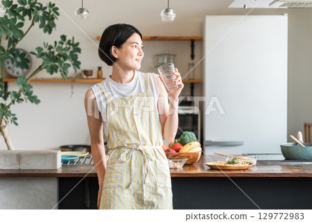 A young Asian woman wearing an apron hydrating in the kitchen 129772983