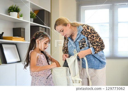 Mother trying to help her daughter pack a school backpack while the girl shows hesitation and reluctance Mother trying to help her daughter pack a school backpack while the girl shows hesitation and reluctance 129773062