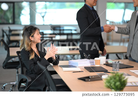 Two businessmen shaking hands in a modern office while a female colleague claps in support 129773095