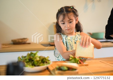 Little girl making a sandwich for breakfast, carefully placing bread over fresh vegetables 129773132