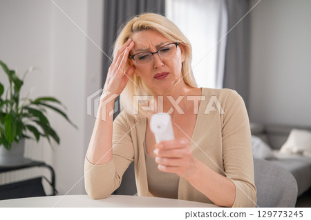Worried blonde woman in beige clothes holding thermometer, experiencing high fever and poor health in modern apartment living room 129773245