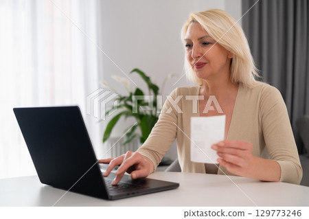 Middle aged blonde in glasses happily working on laptop at home, holding receipt and organizing family finances, modern lifestyle scene Middle aged blonde in glasses happily working on laptop at home, holding receipt and organizing family finances, modern lifestyle scene 129773246