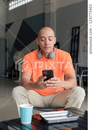 Bald Hispanic man with coffee using phone sending sms in cafe restaurant wearing orange polo shirt 129773403