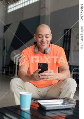 Happy bald Hispanic man with coffee using phone in cafe restaurant wearing orange polo shirt 129773404