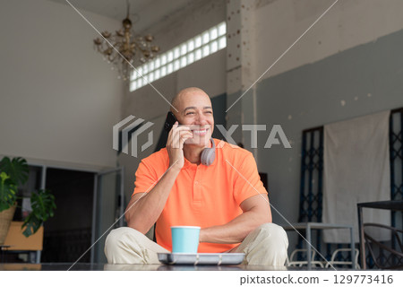 Happy man with coffee talking on phone in cafe restaurant wearing orange polo shirt 129773416