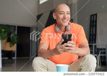 Happy Bald Hispanic man with coffee using phone in cafe and looking away smiling 129773422