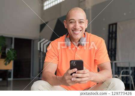 Smiling bald man texting with mobile phone while sitting in coffee shop looking at camera 129773426
