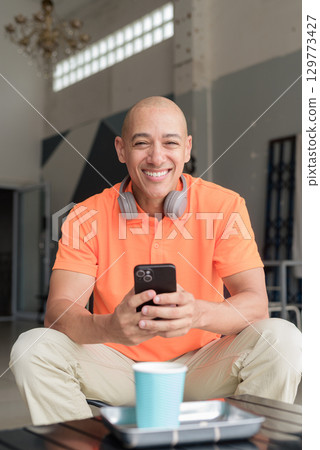 Happy bald man with mobile phone while sitting in coffee shop looking at camera vertical shot Happy bald man with mobile phone while sitting in coffee shop looking at camera vertical shot 129773427