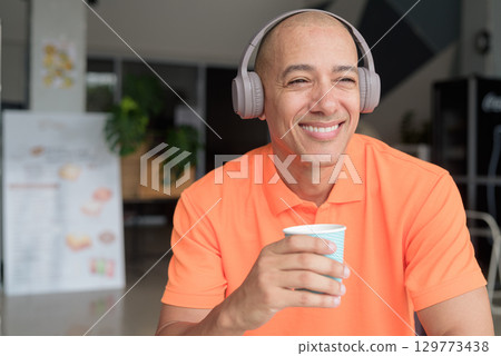 Happy man listening music with headphones while holding coffee cup and looking away 129773438