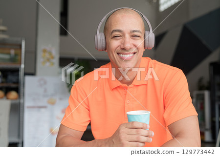 Smiling man listening music with headphones while holding coffee cup looking at camera Smiling man listening music with headphones while holding coffee cup looking at camera 129773442