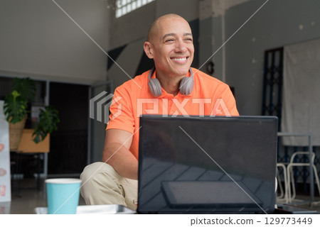 Bald Hispanic man sitting and using laptop computer in coffee shop restaurant smiling and thinking 129773449