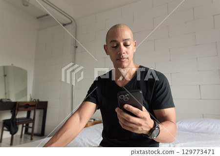 Bald Hispanic man sitting on bed at home using phone 129773471