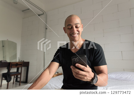 Bald Hispanic man sitting on bed at home using phone while texting and smiling 129773472