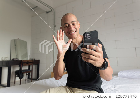 Bald Hispanic man sitting on bed at home using phone having video call waving hand 129773476