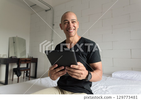 Bald Hispanic man sitting on bed at home using digital tablet computer 129773481