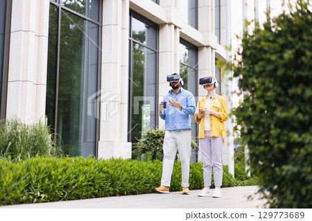 A man and a woman wearing VR headsets are walking outside a modern building. They are both exploring virtual reality. 129773689