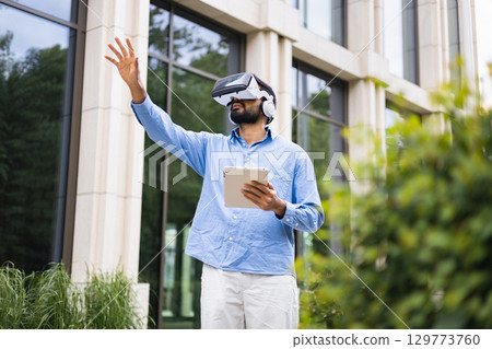 A man in a VR headset outside using a tablet, reaching out. Modern technology outdoors. A man in a VR headset outside using a tablet, reaching out. Modern technology outdoors. 129773760
