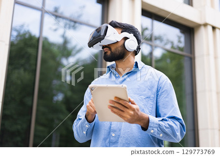 A man wearing a VR headset and headphones, holding a tablet outdoors near a building with large windows. 129773769