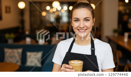 Friendly female barista in a black apron smiling and holding a coffee latte in a cafe Friendly female barista in a black apron smiling and holding a coffee latte in a cafe 129774448