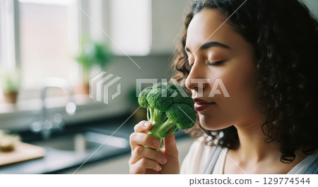 Young woman savoring fresh green broccoli floret in a bright kitchen setting 129774544