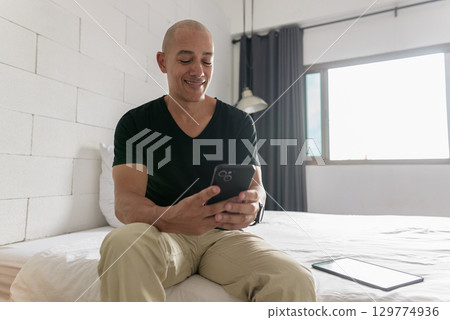Bald Hispanic man sitting on bed at home using phone Bald Hispanic man sitting on bed at home using phone 129774936