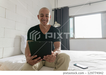 Hispanic bald happy man sitting on bed while using digital tablet computer 129774942
