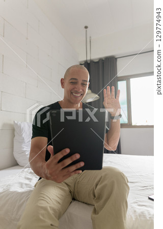 Hispanic bald man sitting on bed while using digital tablet computer 129774943