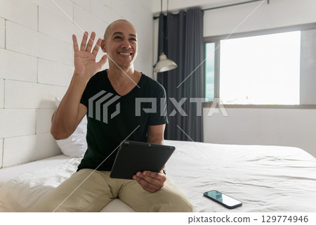 Hispanic bald man sitting on bed while using digital tablet computer while smiling and waving hand 129774946