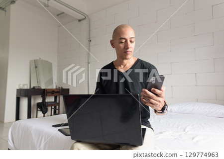 Remote work from home concept bald Hispanic man sitting on bed using laptop computer and phone Remote work from home concept bald Hispanic man sitting on bed using laptop computer and phone 129774963