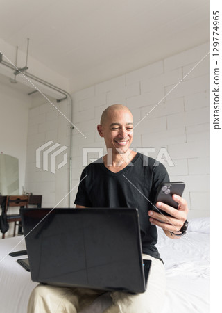 Remote work from home concept bald Hispanic man sitting on bed using laptop computer and phone 129774965