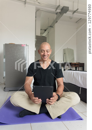 Man sitting on yoga mat doing meditation and wellness exercise at home and using tablet computer 129774980