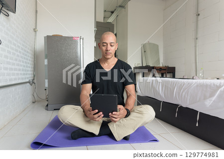 Man sitting on yoga mat doing meditation and wellness exercise at home and using tablet computer 129774981