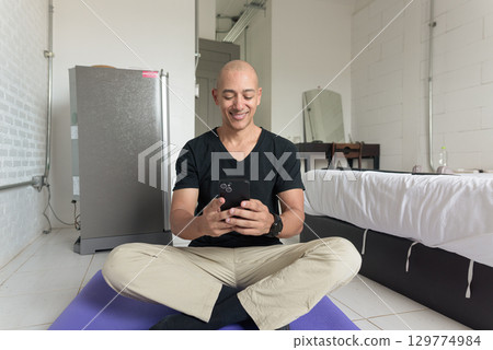 Man sitting on yoga mat doing meditation and wellness exercise at home while using phone 129774984