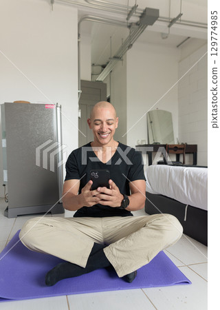 Man sitting on yoga mat doing meditation and wellness exercise at home while using phone 129774985