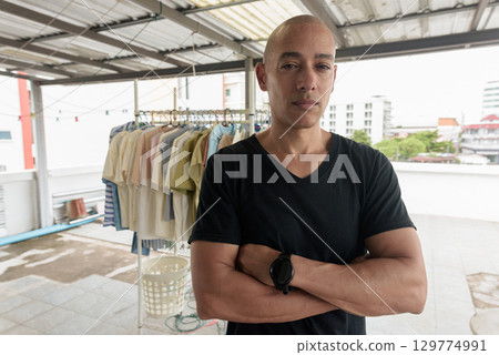 Mature bald Hispanic man with arms crossed in front of drying laundry outdoors Mature bald Hispanic man with arms crossed in front of drying laundry outdoors 129774991
