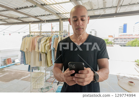 Hispanic bald man using phone while doing laundry outdoors during day 129774994