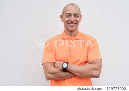 Bald Italian man in orange polo shirt studio portrait against white wall with arms crossed smiling 129775000