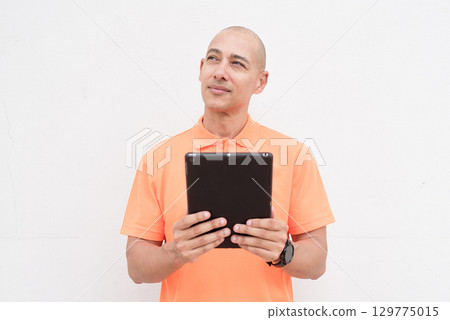 Bald Italian man in orange polo shirt looking up and planning while using digital tablet computer Bald Italian man in orange polo shirt looking up and planning while using digital tablet computer 129775015