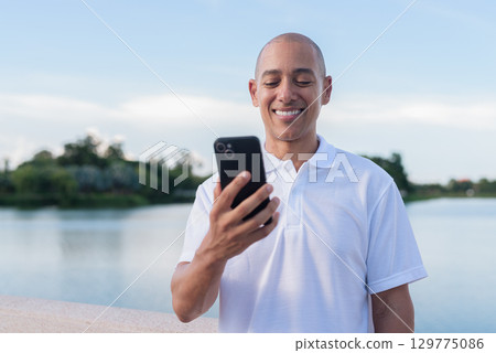 Confident bald man in white polo shirt using phone by lake in serene summer outdoor portrait Confident bald man in white polo shirt using phone by lake in serene summer outdoor portrait 129775086