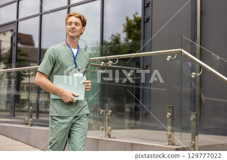 Ginger doctor in lab coat walking by glass clinic building holding medical folder with clinical history 129777022