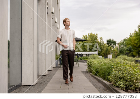 Young male with ginger hair walking outdoors holding closed laptop near modern building 129777058