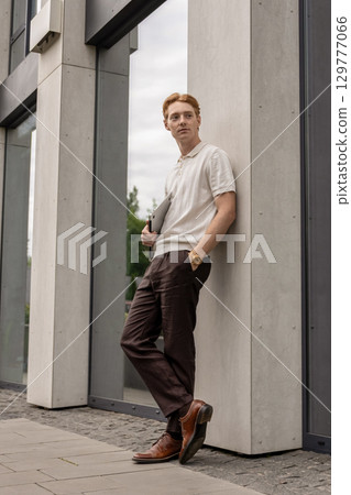 Young male with ginger hair pausing near street wall holding laptop outdoors 129777066