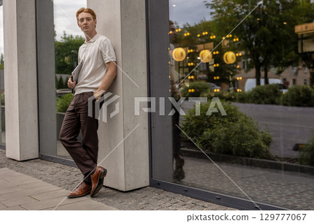 Ginger guy standing still near neutral wall holding laptop and focusing on something in distance 129777067