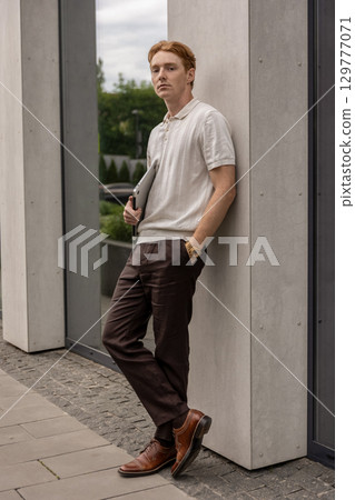 Young ginger male standing near wall holding laptop on street 129777071