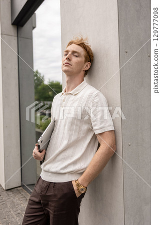 Ginger-haired man pausing on the street with laptop in his hand near urban building Ginger-haired man pausing on the street with laptop in his hand near urban building 129777098